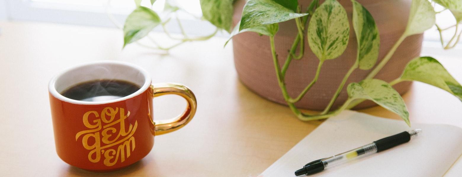 mug of tea on a table next to a plant and notebook with pen indoors